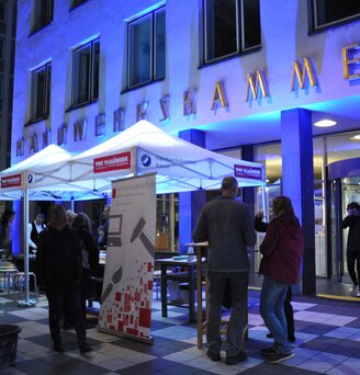 White 3x3m event gazebo customised with Handwerkskammer logo with blue lights at night sheltering people and tables at an event