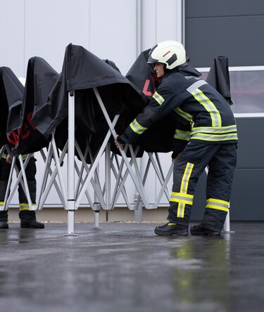German firefighter in uniform setting up a black 3x3 m Ecotent pop-up canopy on a rainy day.