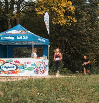 The blue and white event tent serves as a supply station for a marathon. 2 runners are passing the event tent.